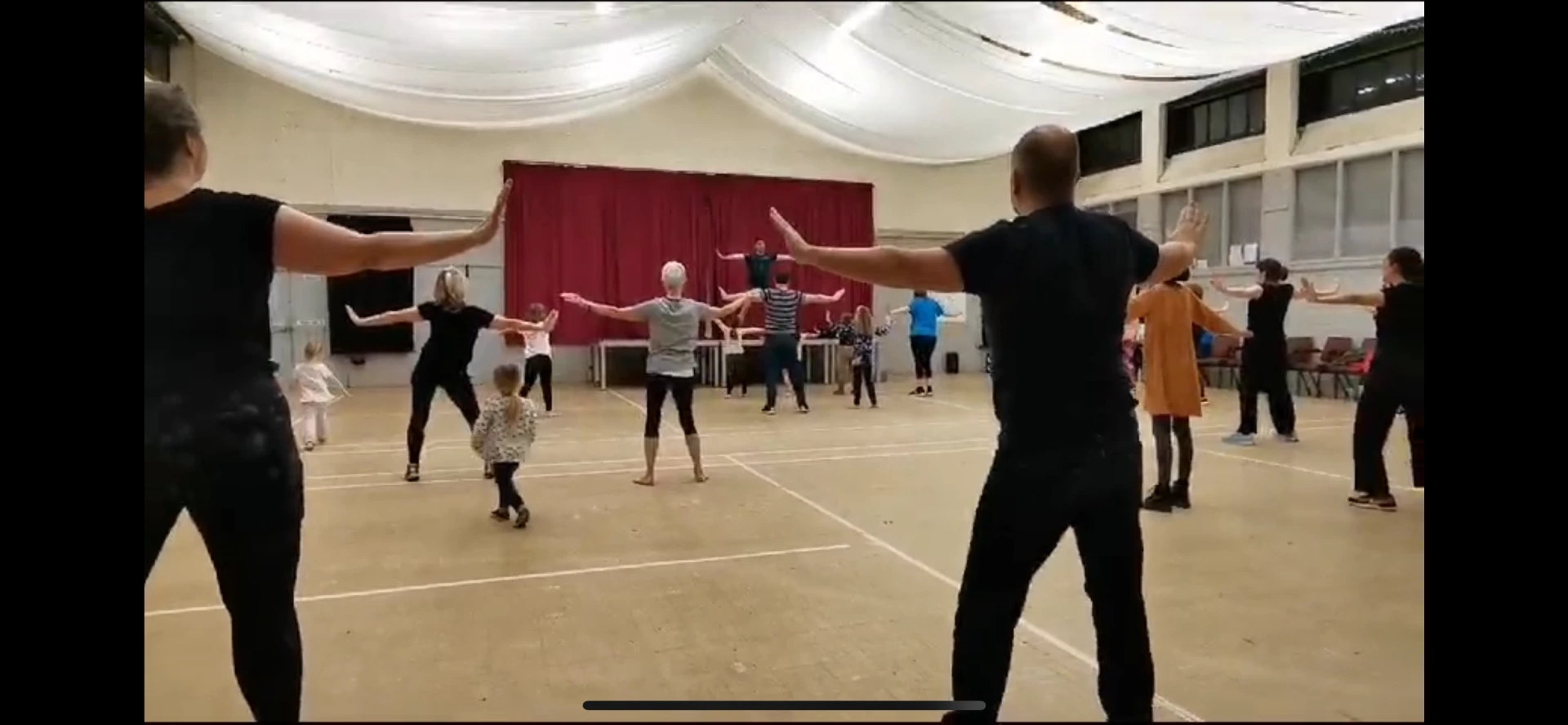 Full dance class in a village hall following Jason with arms outstretched